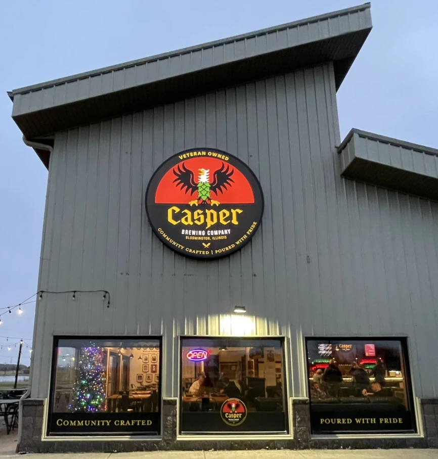 The Casper Brewing Company taproom at dusk. The round lit sign hangs above three window graphic panels.