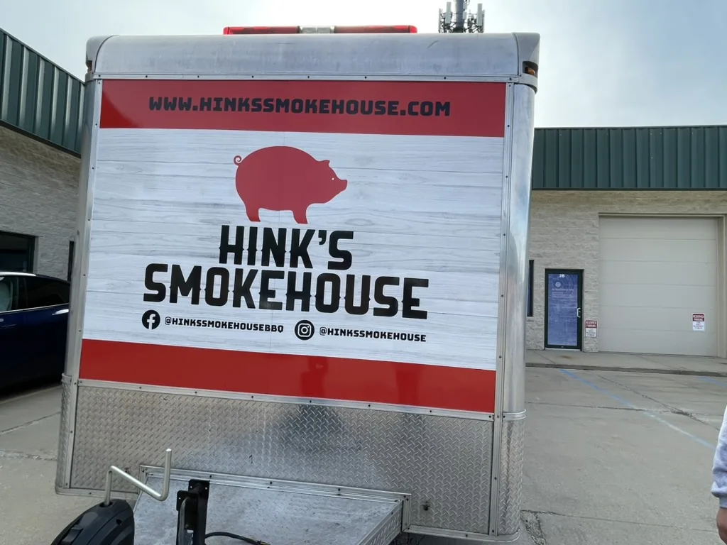 Rear of the Hink's Smokehouse BBQ trailer with pig mark, web address, and social handles, Bloomington, IL.
