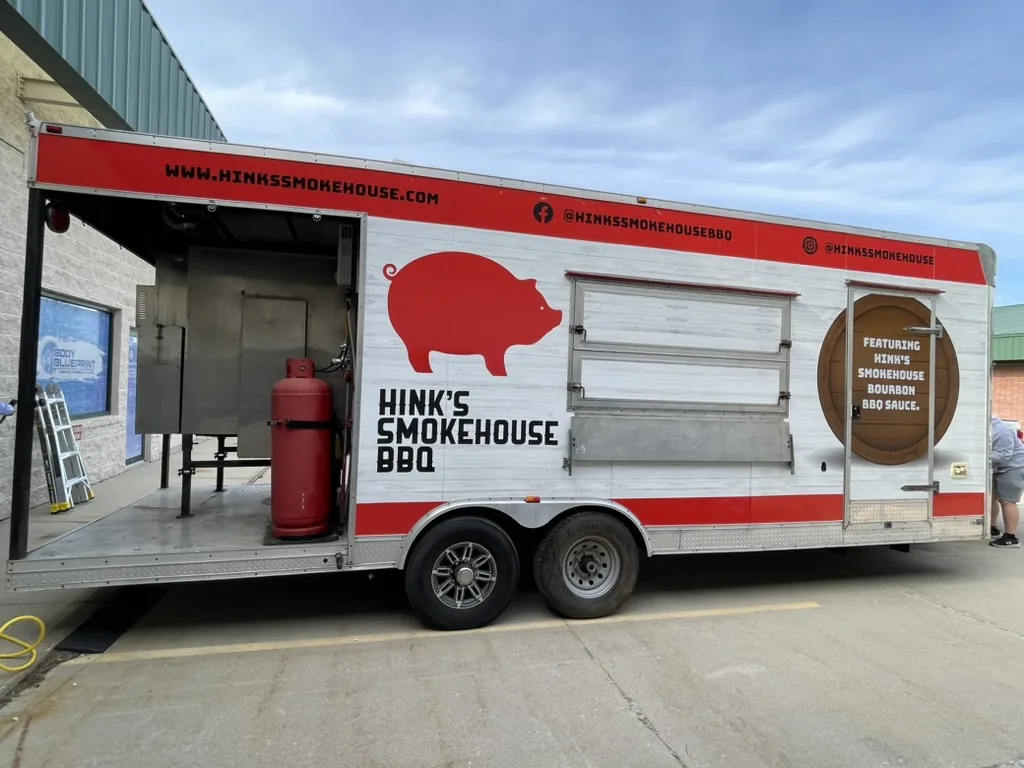 Serving side of the Hink's Smokehouse trailer with wrap graphics running continuously across serving windows and a bourbon barrel graphic on the entry door, Bloomington, IL.