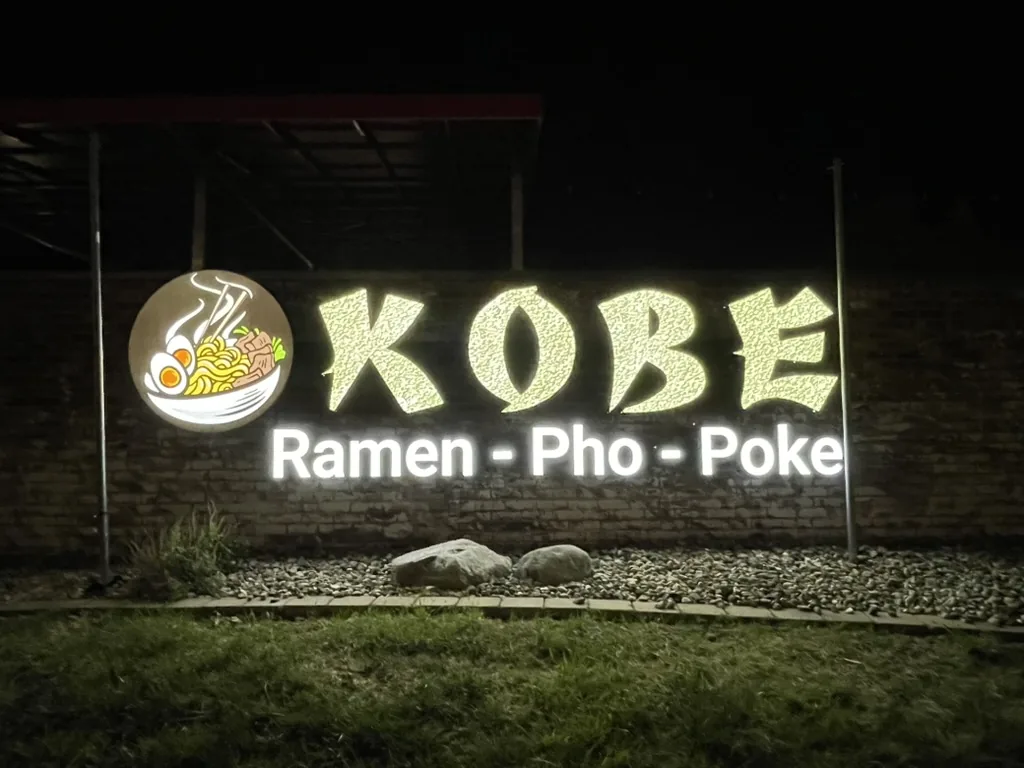 Large lighted Kobe Ramen Pho Poke wall sign with an illuminated ramen bowl logo on a brick wall at night in Bloomington, IL.