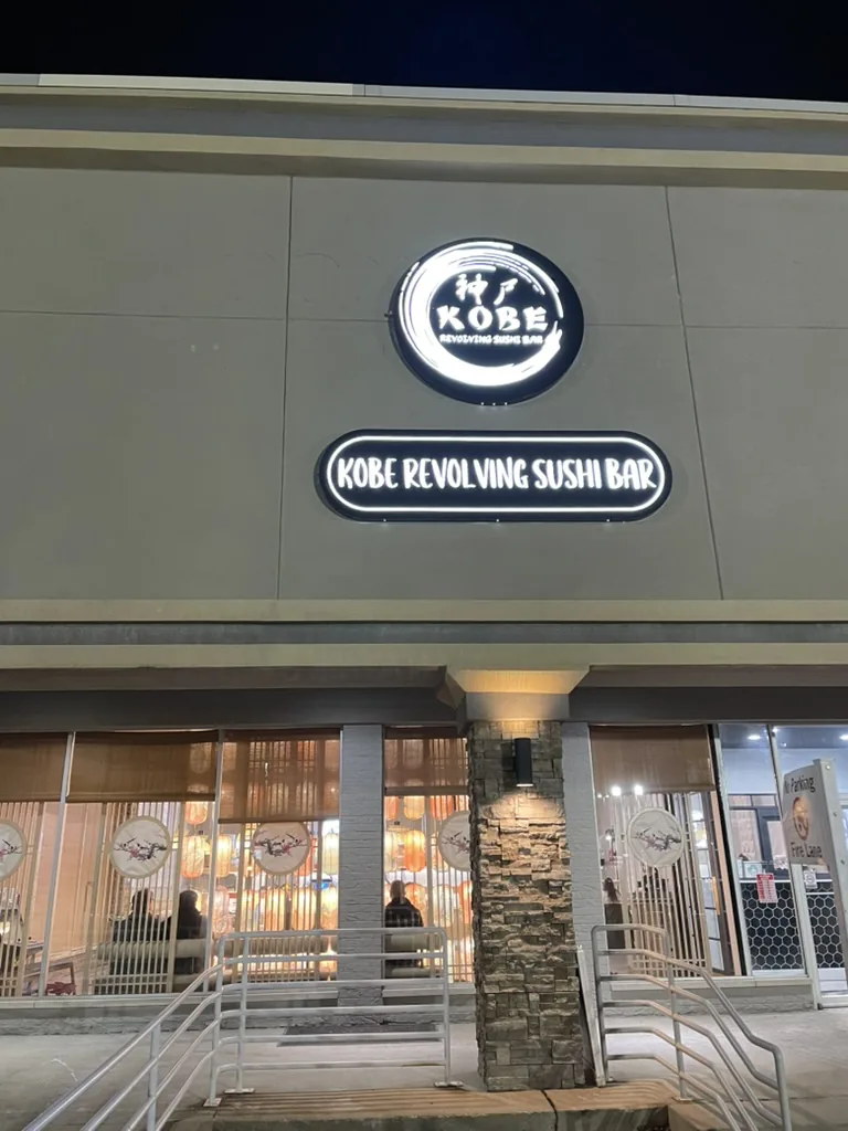 Lighted circular logo sign and oval lightbox for Kobe Revolving Sushi Bar above the storefront at night in Bloomington, IL.