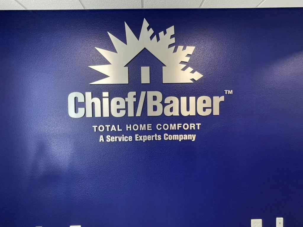 Dimensional letters and a logo on a painted blue interior wall. The logo shows a house with a sun on one side and a snowflake on the other. The letters read Chief/Bauer, Total Home Comfort, A Service Experts Company.