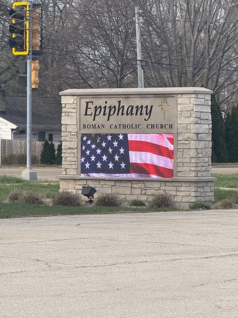 The monument seen from across the road, with the message center running a full-color image. A traffic signal and overhead lines cross above it, houses stand behind, and the church parking lot runs into the foreground.