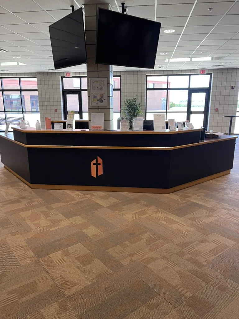 The six-sided information desk wrapped in matte black, with the orange Grace Church shield on the front and two screens hung above it.