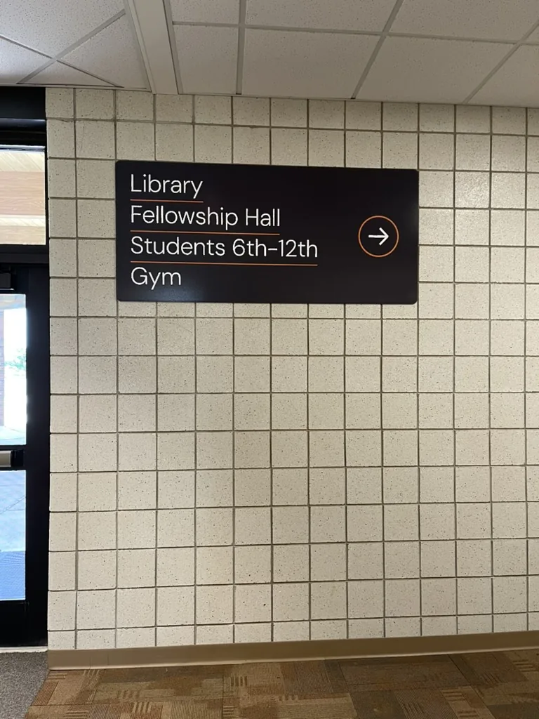 A black wayfinding panel on a tile wall listing Library, Fellowship Hall, Students 6th to 12th, and Gym, with an orange arrow pointing right.